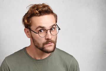 Portrait of young man with specific appearance looks through round glasses, listens attentively to...