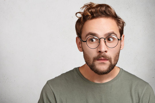 Headshot of attractive stylish youngster or male wonk wears round spectacles, looks curious aside, tries to overhear somethinng important. Caucasian bearded young man with guarded look, worries