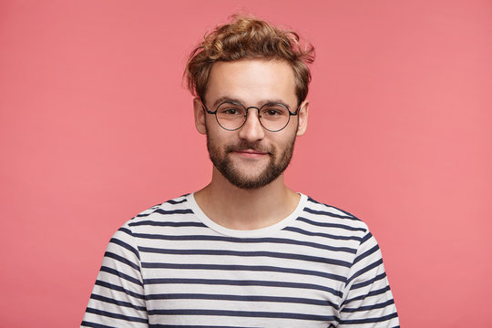 Indoor shot of pleasant looking bearded hipster guy wears spectacles and striped t shirt, looks directly into camera, isolated over pink studio background. Male student spends weekends at home alone