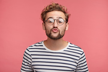 Funny male with curly trendy hairstyle, rounds lips as going to kiss someone, looks in anticipation, isolated over pink background. Fashionable bearded man epresses positive emotions, makes grimace