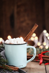 Enamel cup of hot cocoa with mini marshmallows and cinnamon bark. Pine boughs and gray scarf...