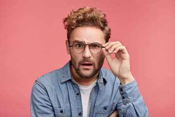 Scrupulous stylish man in denim shirt and spectacles looks with shocked and stupefied expression,...