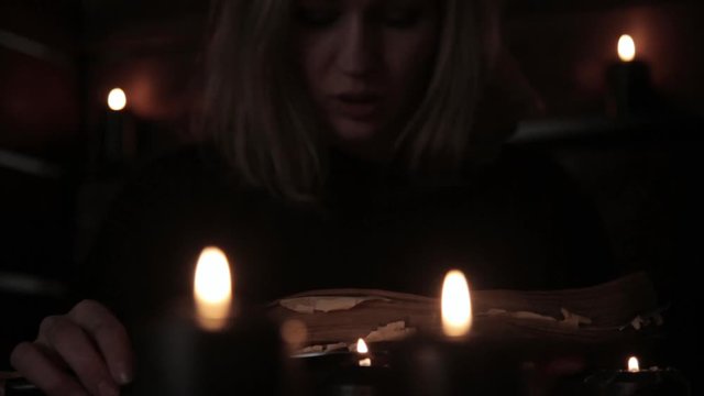 witch holds a knife and reading spell from old magic book lit by black candles
