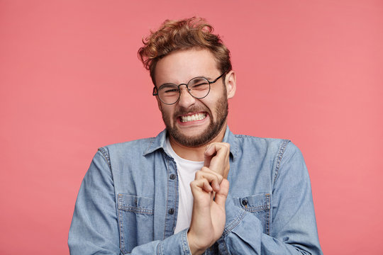 Stylish young male clenches teeth, has digusting expression, refuses do something, makes grimace, isolated over pink background. Dissatisfied youngster in glasses feels averse to do something