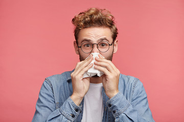 Indoor shot of bearded man has running nose, rubs nose with handkerchief, being ill, caught cold after long walk outdoors on rainy weather. Unhealthy allergic male sneezes isolated over pink wall