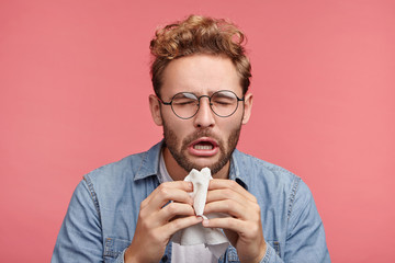 Portrait of ill or sick bearded male sneezes and coughs, uses handkerchief, rubs nose, being allergic to animals. Man has running nose, caught cold when had walk outdoors in winter or autumn - obrazy, fototapety, plakaty