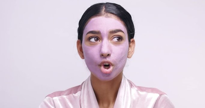 Pretty Middle Eastern young female model in pink clay cleaning mask making funny faces isolated on white