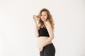 Fototapeta premium Young attractive cheerful pregnant women with wavy light hair in sport comfy outfit smiling, holding hair with hand, touching belly, looking in camera posing for article about successful mothers.