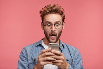 Emotional bug eyed handsome man has trendy hairdo, holds modern mobile phone, being shocked to recieve message from partner, completely forgets about their meeting, has no time for preparation
