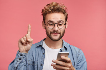 Young curly stylish hipster guy dressed in jean shirt, holds smart phone, reads something...