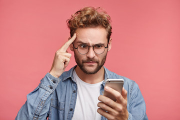 Portrait of serious male keeps finger on temples reads information on mobile phone, upset to find out about raising sales. Stressful puzzled male model uses smart phone to find out news from internet
