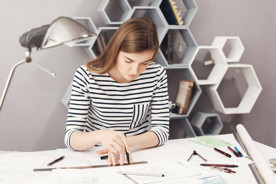 Portrait of serious young good-looking female architect sitting at her workplace, making drawings with pencil and ruler, trying not to make a mistake in blueprints.