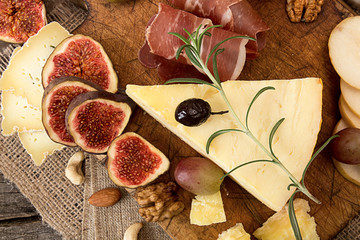 cheese plate with pieces moldy cheese, prosciutto, pickled plums, olives, grapes, figs, honey, pear,  walnuts, almonds, cashews on old wooden background. Rustic style.