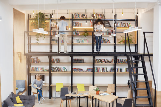 Phto of big modern univesity library. Blonde girl sitting on chear looking in window with dreamy face expression. Two young people standing near bookshelves, reading books.