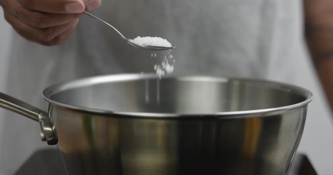 Chef in gray apron adds sea salt to a stainless steel pot of boiling water