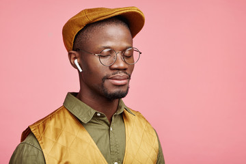 Sideways portrait of serious relaxed dark skinned fashionable male wears trendy cap, spectacles, listens to restful music in wireless earphones, enjoys calm atmosphere, isolate over pink wall