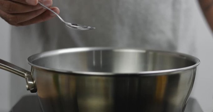 Chef in gray apron adds sea salt to a stainless steel pot of boiling water and mixes with a wooden spatula
