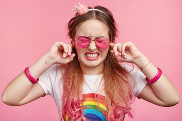 Young frustrated stylish female likes pink colour, blocks ears as hears loud music on party, has headache, clenches teeth, wants silence, isolated over pink background. Cute lovely girl plugs ears