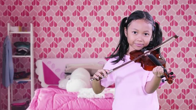 Adorable little girl with pigtail hair, playing a violin in the bedroom with pink color decoration at home