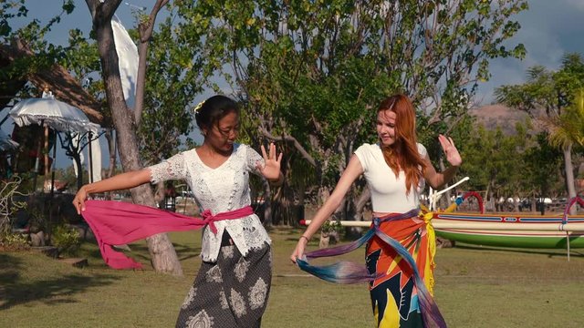 Balinese girl teaching caucasian woman traditional dance moves with sash, wearing sarong outdoors on green lawn. Shot with Sony a7s and Atomos Ninja Flame on sunny summer day.