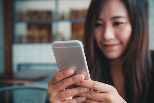 Closeup image of a beautiful Asian woman with smiley face holding and using smart phone  in modern cafe