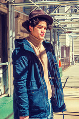 American Man traveling, walking through sidewalk bridge in New York. Wearing blue jacket with hood, knitted turtleneck sweater, Fedora hat, guy with freckle face, curly hair, looking at you under sun