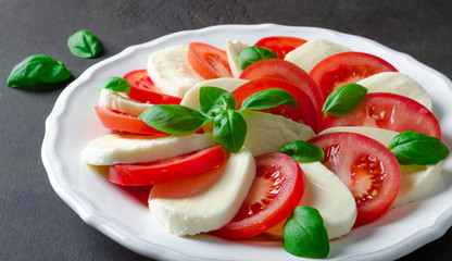 Sliced tomatoes and mozzarella on a white plate, dark stone background. Closeup, selective focus