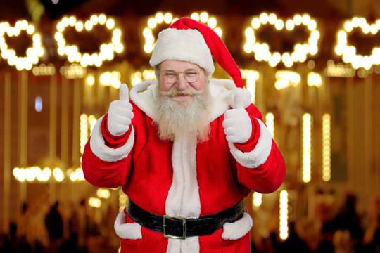 Old Santa Claus giving thumbs up with both hands. Positive Santa Claus giving thumb up gesture on New Year lights background. Realistic Santa Claus on blurred background.