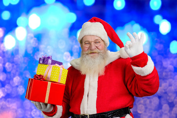Santa Claus with Christmas gifts. Realistic Santa Claus holding boxes with presents and showing sign ok, blue shimmering background.