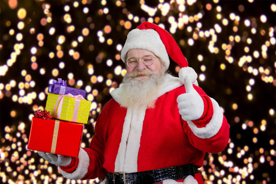 Santa with gifts showing thumb up. Santa Claus holding a pile of gifts boxes and giving thumb up on New Year lights background.