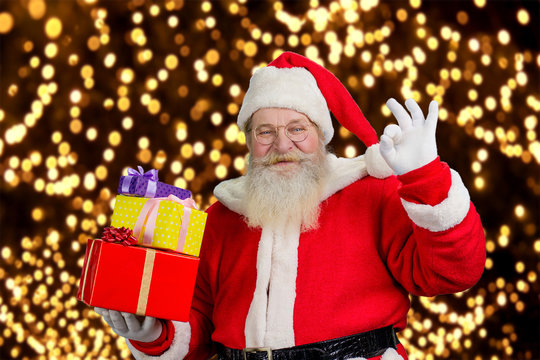 Happy Santa on New Year lights background. Smiling male Santa Claus holding pile of Christmas presents and showing okey sign. Santa Claus with gift boxes.