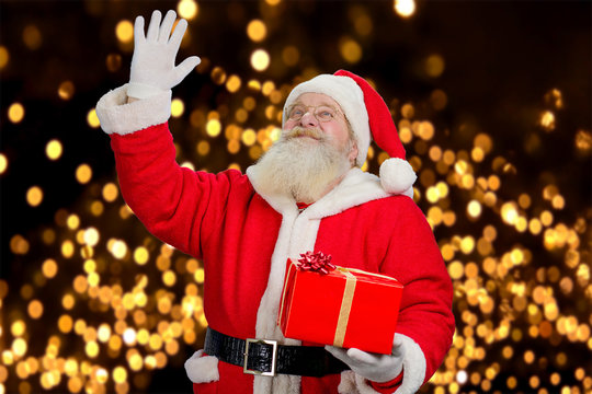 Santa Claus waving with hand. Portrait of realistic Santa Claus with Christmas gift waving with hand on New Year lights background. Christmas time and presents.