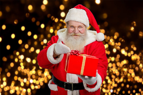Happy Santa Claus offering present. Kind Santa Claus with red gift box on festive lights background.