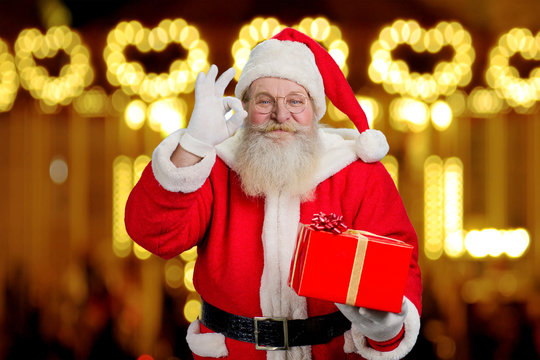 Smiling Santa Claus showing okey sign. Santa Claus with real beard holding Christmas present and making okey gesture with fingers on blurred background.