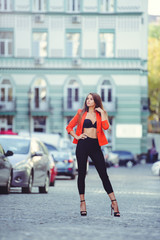 Fashionable look, hot day model of a young woman walking in the city, wearing a red jacket and black pants, blond hair outdoors over the city warm background