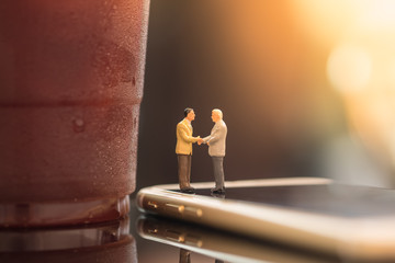 Fototapeta premium Coffee, Time and Business cooperation concept. Two businessman miniature figures meet and hand shake on smart phone and close to plastic cup take away of iced black coffee.