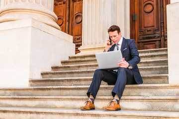 American Businessman traveling, working in New York, dressing in black suit, leather shoes, sitting on stairs outside office, working on laptop computer, calling on phone. Instagram filtered effect..