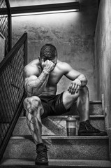 Bodybuilder resting after hard workout. He`s seating on the stairs in the gym with shaker full of fitness supplement.