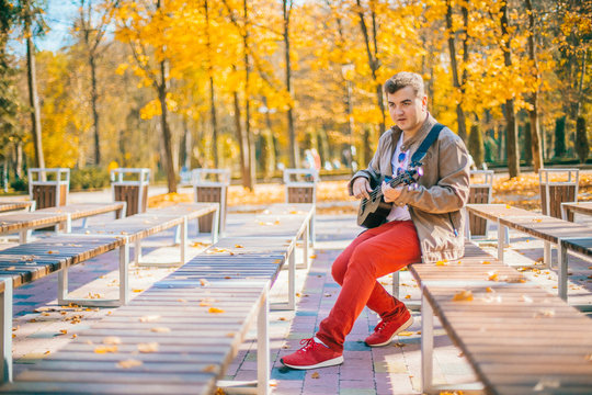 Young man are playing bass ukulele on the park autumn park near lake. Basking guy at the street. Two case for guitars ukulele.