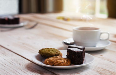 chocolate cookies and brownie in white plate on wood table with coffee cup at morning with sunrise. dessert and coffee concept.