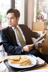 asian business man reading newspaper with morning sandwish in coffee shop