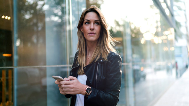 Fashion blogger is using typing messages on the mobile phone while standing on a street background. Young stylish blonde female is reading emails on the smartphone while passing by a glass building.