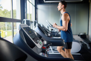 Side view of young man running on treadmill in modern gym by window, focus in foreground on row of...