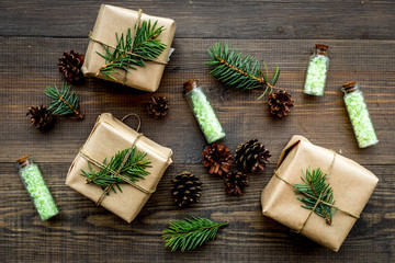 Cosmetics with pine oil. Spa salt in bottles near wrapped boxes, spruce branches, pinecones on wooden background top view