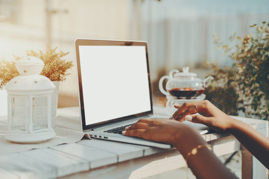 Beautiful and neat hands of African American female with beige manicure, using laptop with white blank screen mock-up in street cafe with glass teapot in background; wooden white table, sunny day