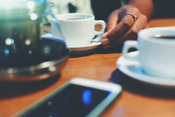Shallow depth of field shooting of neat female hand holding plate with cup on it with delicious coffee on wooden table surrounded by another cup, glass teapot and smartphone in defocused foreground