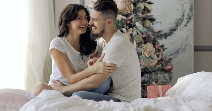 young beautiful couple sitting on a bed next to a Christmas tree, hugging, kissing and laughing
