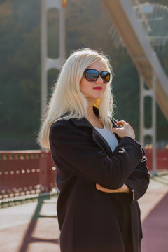 Beauty blonde model posing in black coat wears sunglasses  on a blurred city background