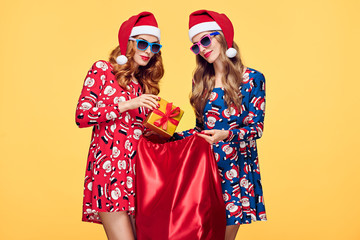 Christmas. Young Woman and Santa Sack with Presents Having Fun Happy Smiling. New Year Fashion. Best Friends Twins in Stylish fashion Red Xmas Holiday Dress on Yellow.Christmas Colorful