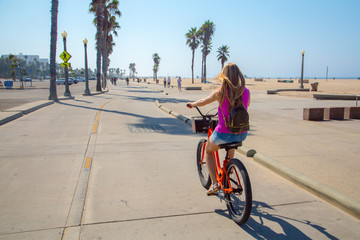 Young beautiful girl riding a bike down the Venice beach in Los Angeles. Californian dreaming. - obrazy, fototapety, plakaty